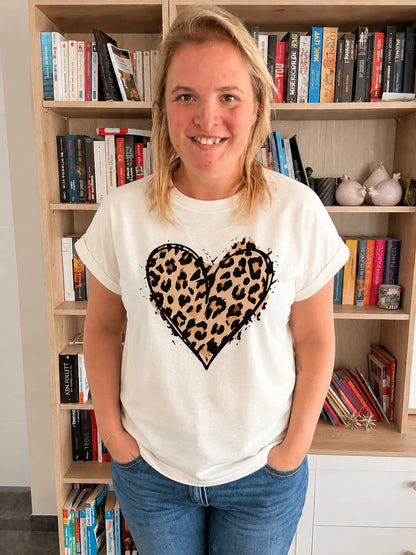 T-shirt blanc femme avec cœur imprimé léopard, col rond et manches courtes, portée par une femme devant une étagère de livres.