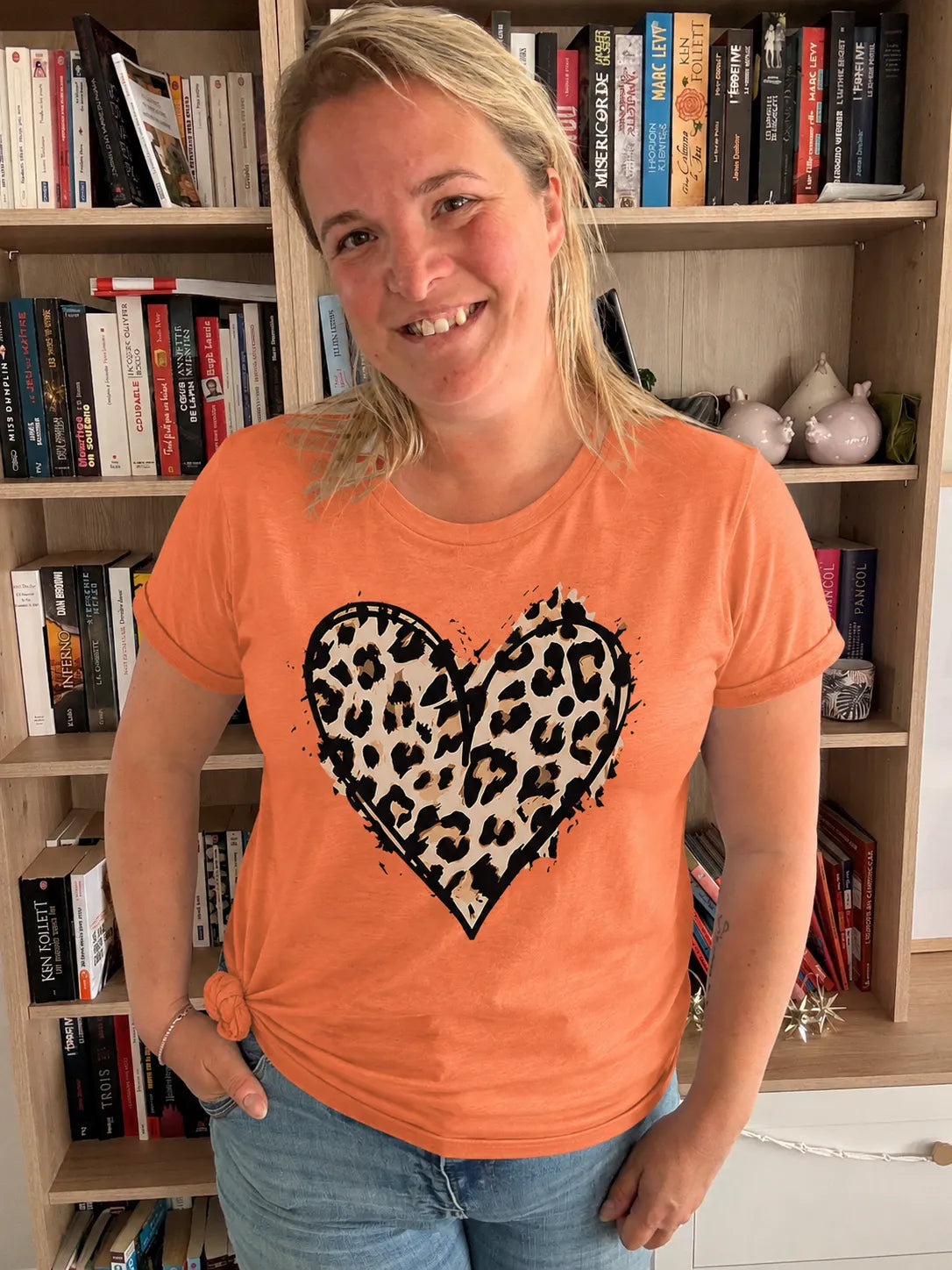 T-shirt femme orange avec imprimé cœur léopard noir et beige, col rond, porté par une femme souriante dans une bibliothèque.