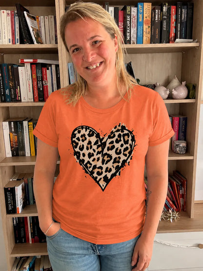T-shirt femme orange avec cœur imprimé léopard noir, porté par une femme souriante dans une pièce avec étagères et livres. Col rond.
