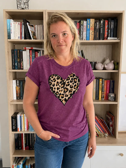 T-shirt femme violet avec cœur imprimé léopard, portée par une femme debout devant une étagère remplie de livres.
