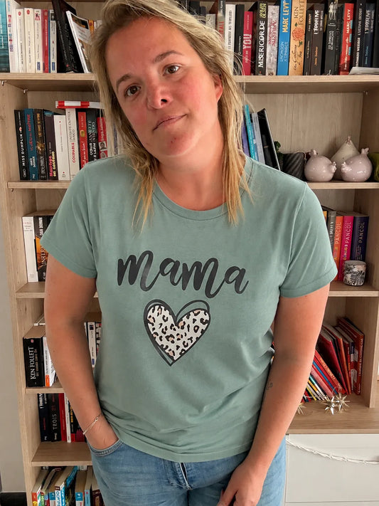 T-shirt vert sauge avec imprimé cœur léopard et texte « Mama » sur une femme debout dans une bibliothèque. Couleur et coupe décontractée.