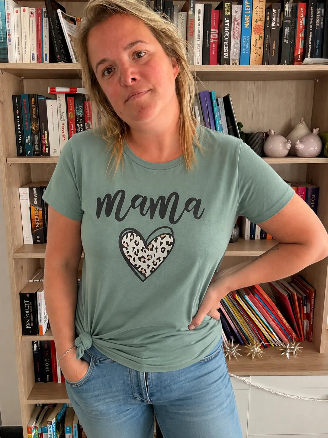 T-Shirt Vert Sauge graphique « Mama » avec cœur léopard, coupe décontractée, portée par une femme debout près d’une bibliothèque.