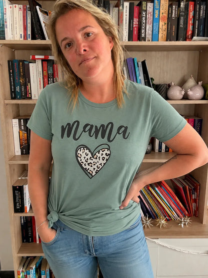 T-Shirt Vert Sauge graphique « Mama » avec cœur léopard, coupe décontractée, portée par une femme debout près d’une bibliothèque.