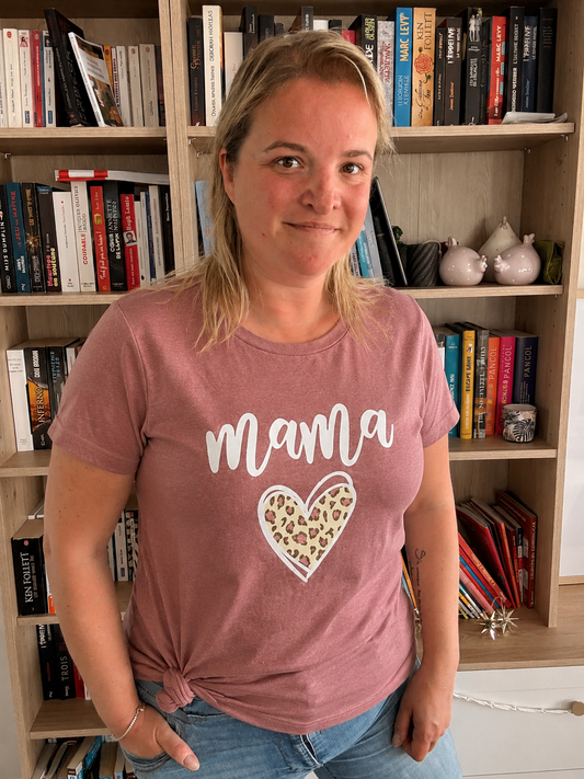T-shirt rose clair femme avec imprimé “mama” et cœur léopard, manches courtes, noué sur le côté, debout devant une bibliothèque.
