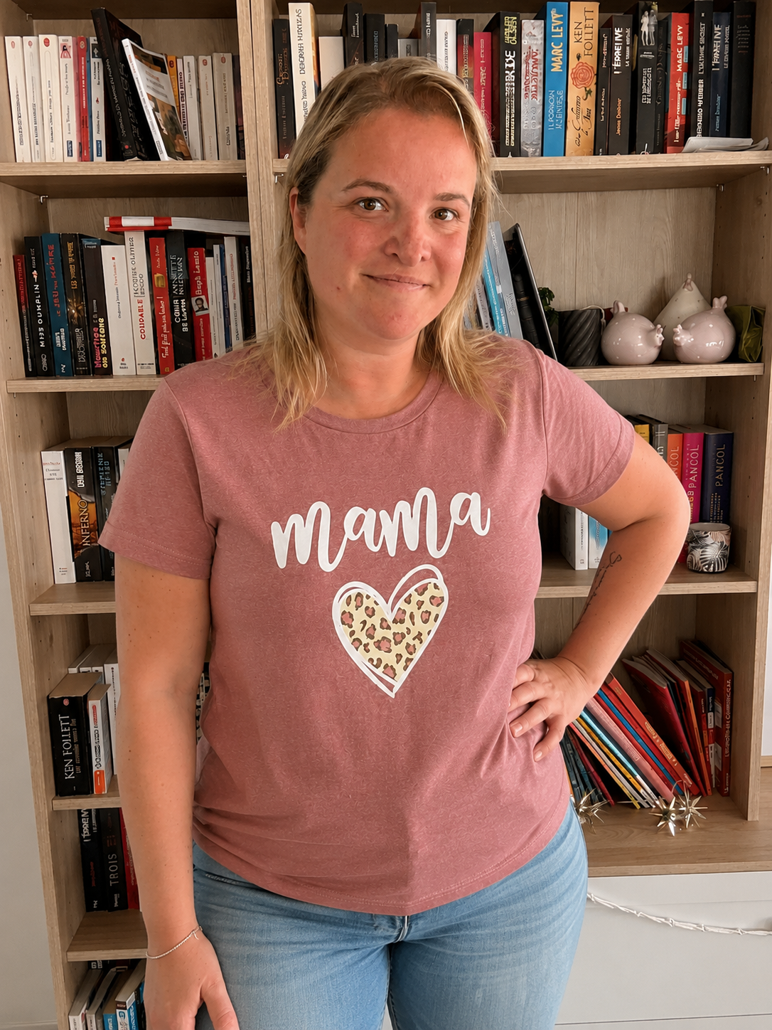 T-shirt rose mauve à manches courtes avec le mot « maman » et cœur léopard imprimé sur le torse, femme debout près d’une bibliothèque.