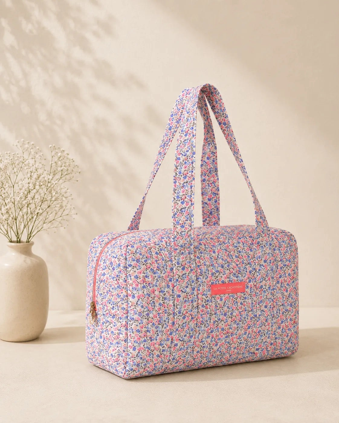 Sac polochon matelassé fleuri imprimé Liberty lilas, rose et bleu avec bandoulières, posé sur une table.
