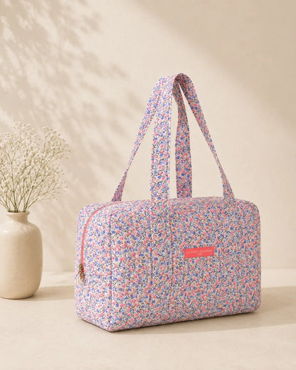 Sac polochon matelassé fleuri imprimé Liberty lilas, rose et bleu avec bandoulières, posé sur une table.