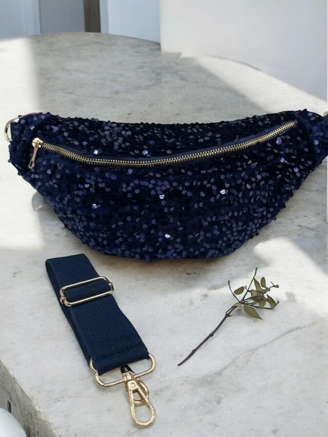 Sac banane bleu marine à paillettes avec bandoulière réglable, zippée dorée et anse rembourrée, accompagnée d'une sangle et d'une petite branche.