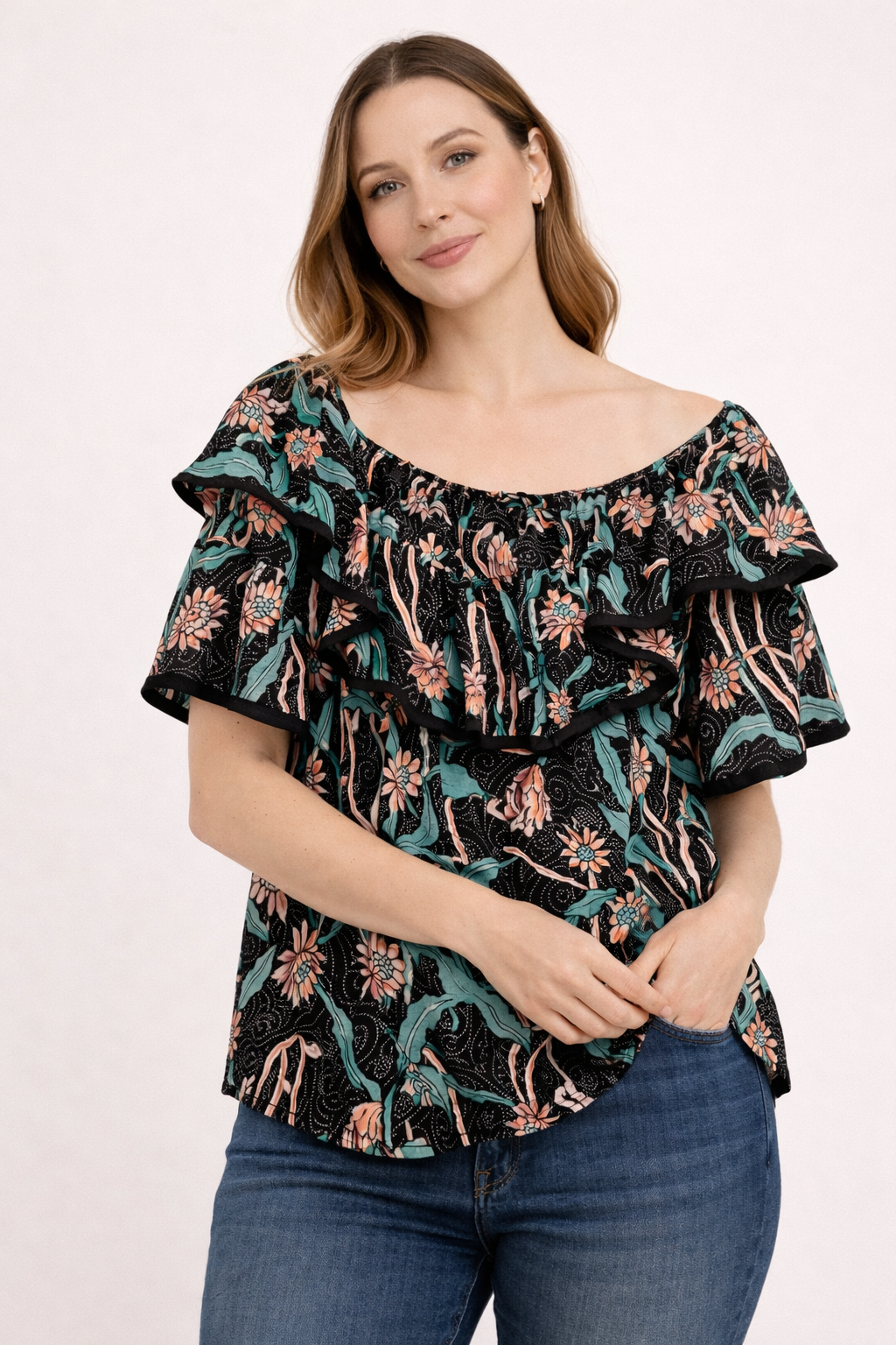 Blouse florale noire à col volanté portée par une femme assise en jeans, motif floral peach et vert sur fond noir.