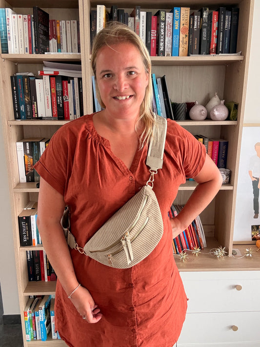 Homme ou femme portant sac banane velours côtelé taupe avec 2 poches et sangle déployée en bandoulière, debout devant une bibliothèque.