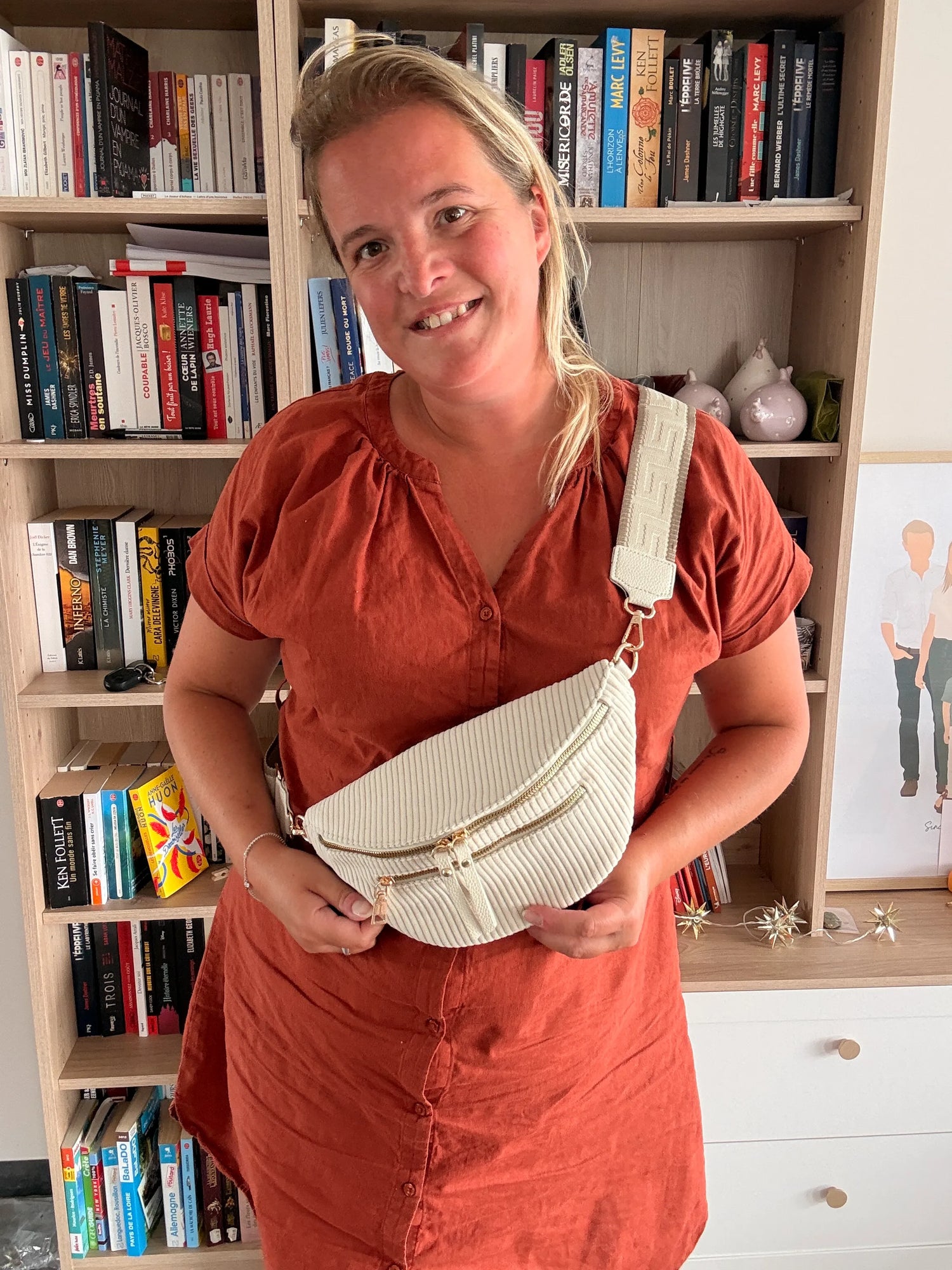 Sac banane velours côtelé beige avec sangle géométrique et zips dorés, portée par une femme en robe orange dans une bibliothèque. Best-seller.