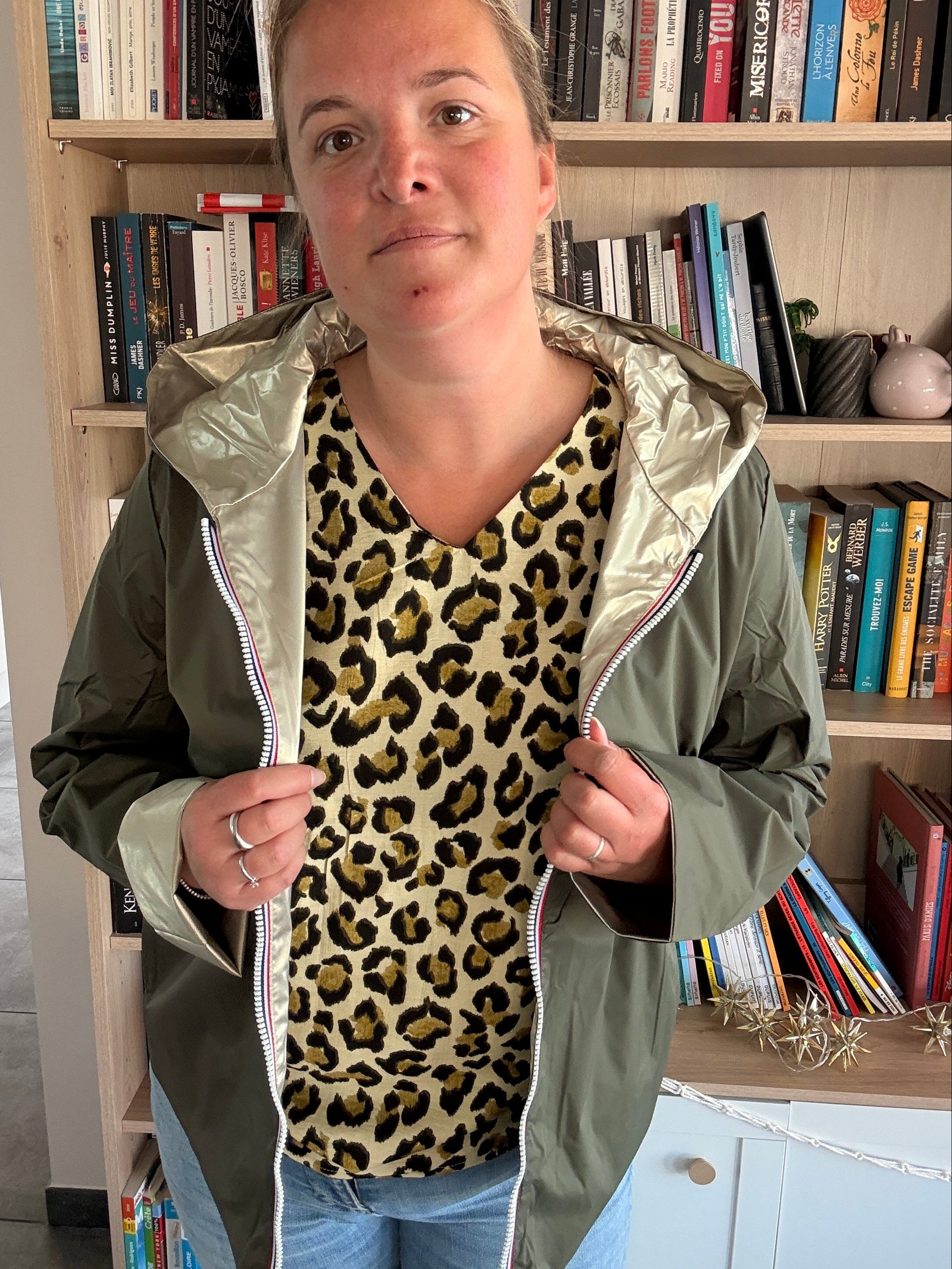 Imperméable réversible kaki/doré porté par une femme en intérieur, veste légère avec capuche, devant bibliothèque. Inclure le mot-clé si pertinent.