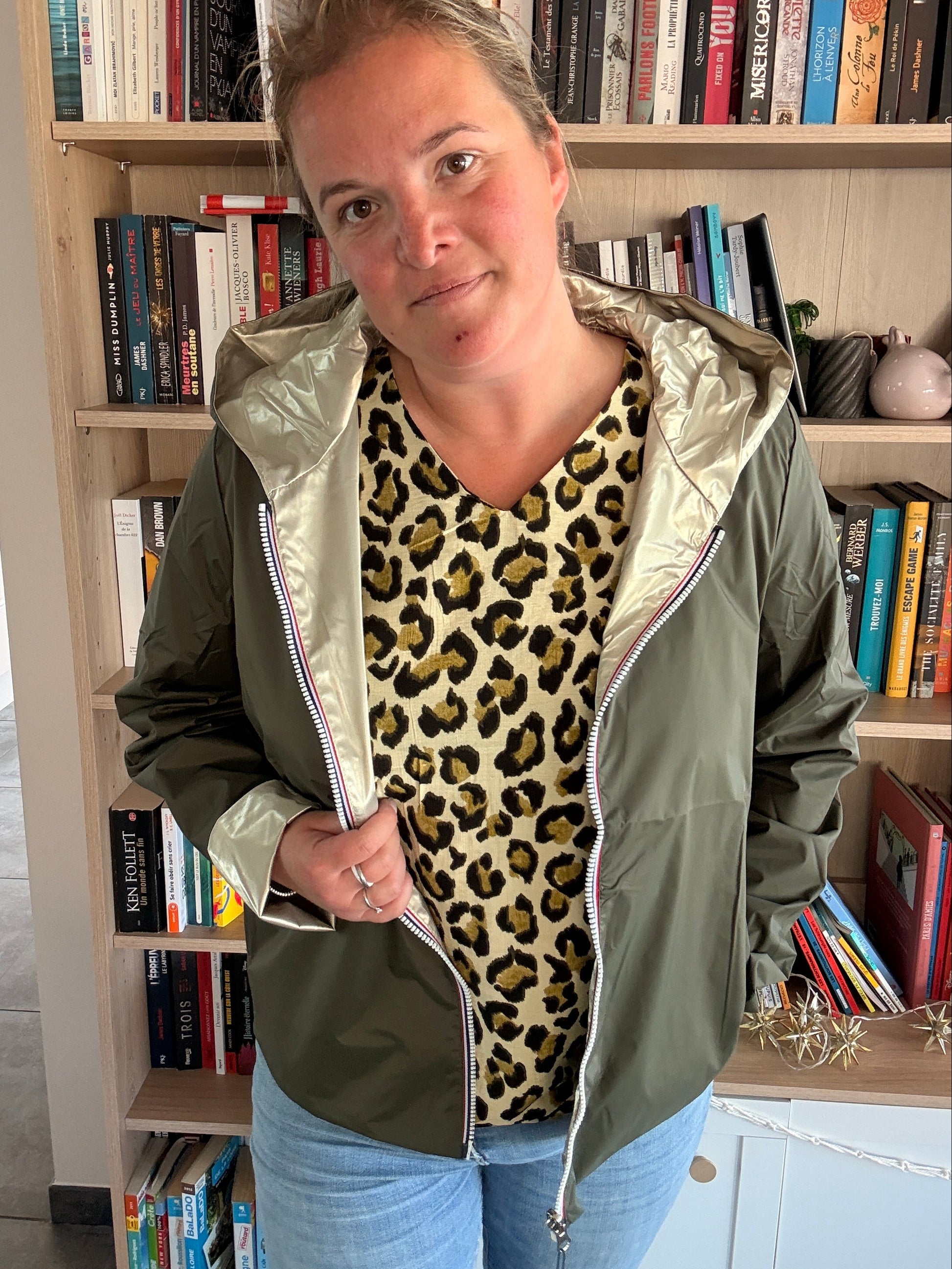 Imperméable réversible court kaki/doré porté par une femme, veste légère ouverte sur un pull léopard et jean, en intérieur.