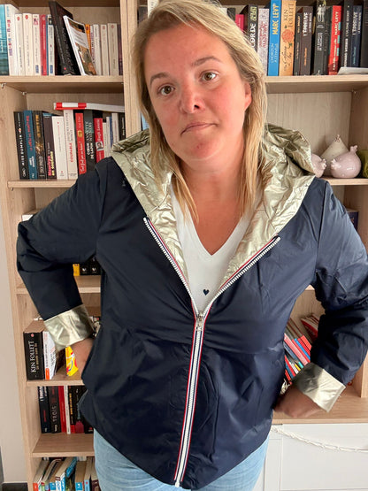 Imperméable réversible court bleu marine doré pour femme, porté par une modèle, avec doublure dorée et zips rouges visibles; fond bibliothèque.
