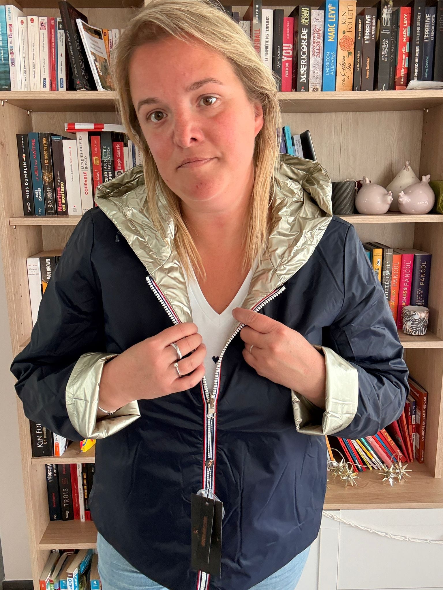 Imperméable réversible court bleu marine doré femme, portée devant une étagère de livres; col et poignets dorés brillants, fermeture éclair argentée, badge suspendu.