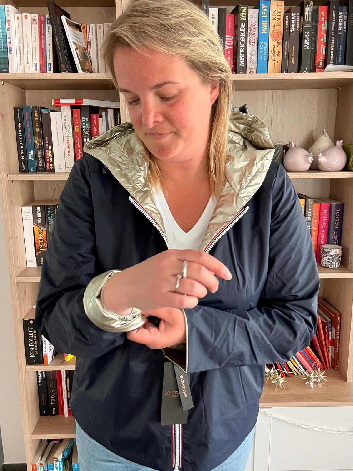Imperméable réversible court bleu marine et doré porté par une femme, montre le poignet et l’étiquette, fonds bibliothèque.