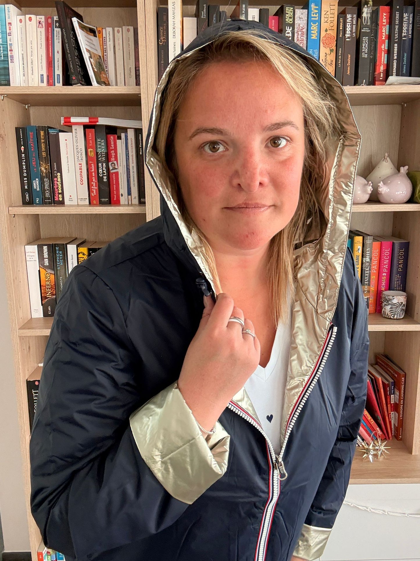 Imperméable réversible court bleu marine doré femme, portée par une femme devant une étagère de livres, capuche dorée et bordures dorées visibles.