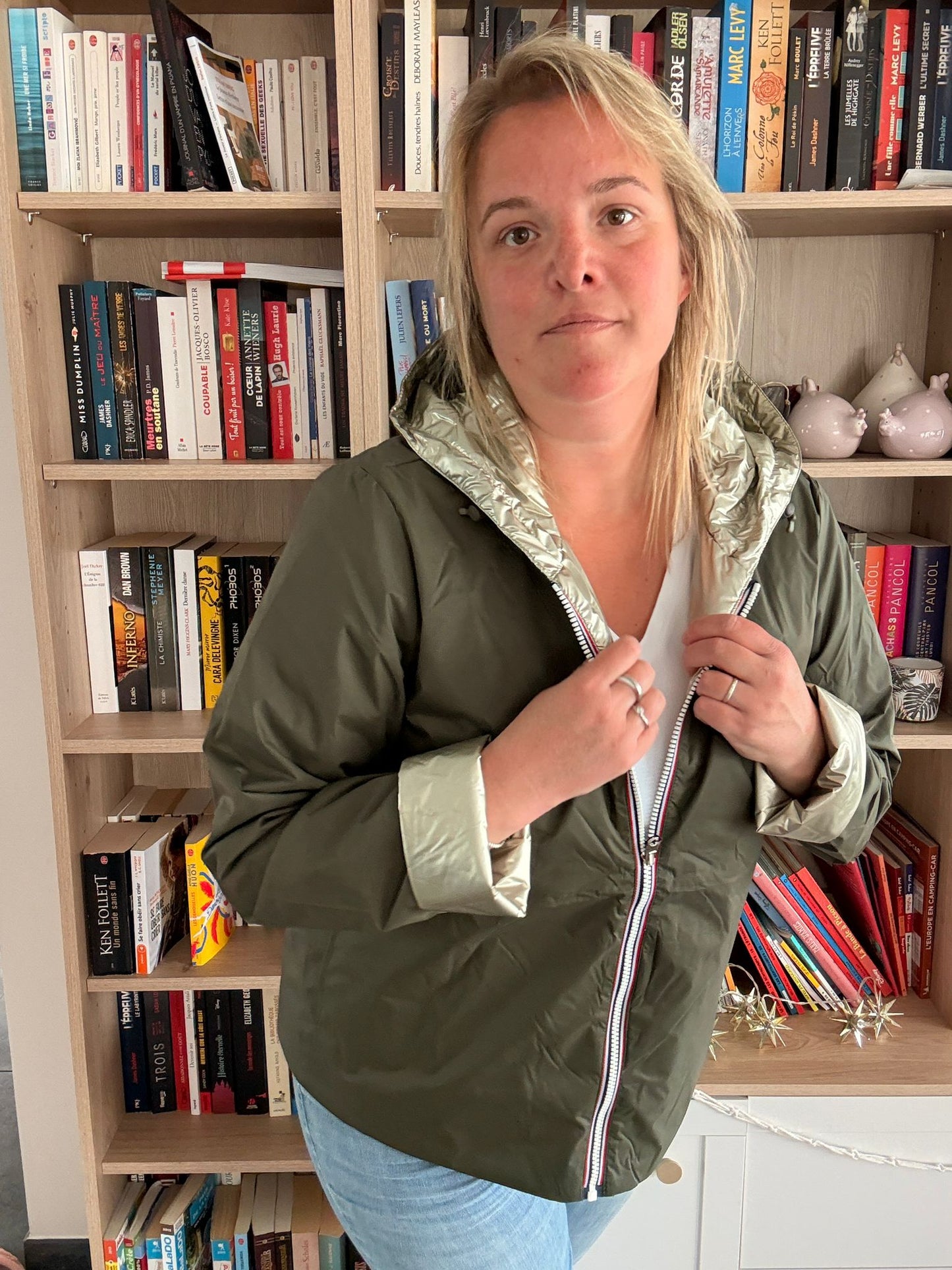 Femme portant un imperméable réversible court kaki doré, debout à côté d'une bibliothèque, en train de tenir les colliers de fermeture zippée sur le devant.