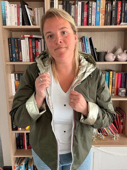 Imperméable réversible court kaki doré pour femme, portée par une femme debout dans une pièce avec étagères de livres; elle tient les pans du manteau, veste mi-saison légère.