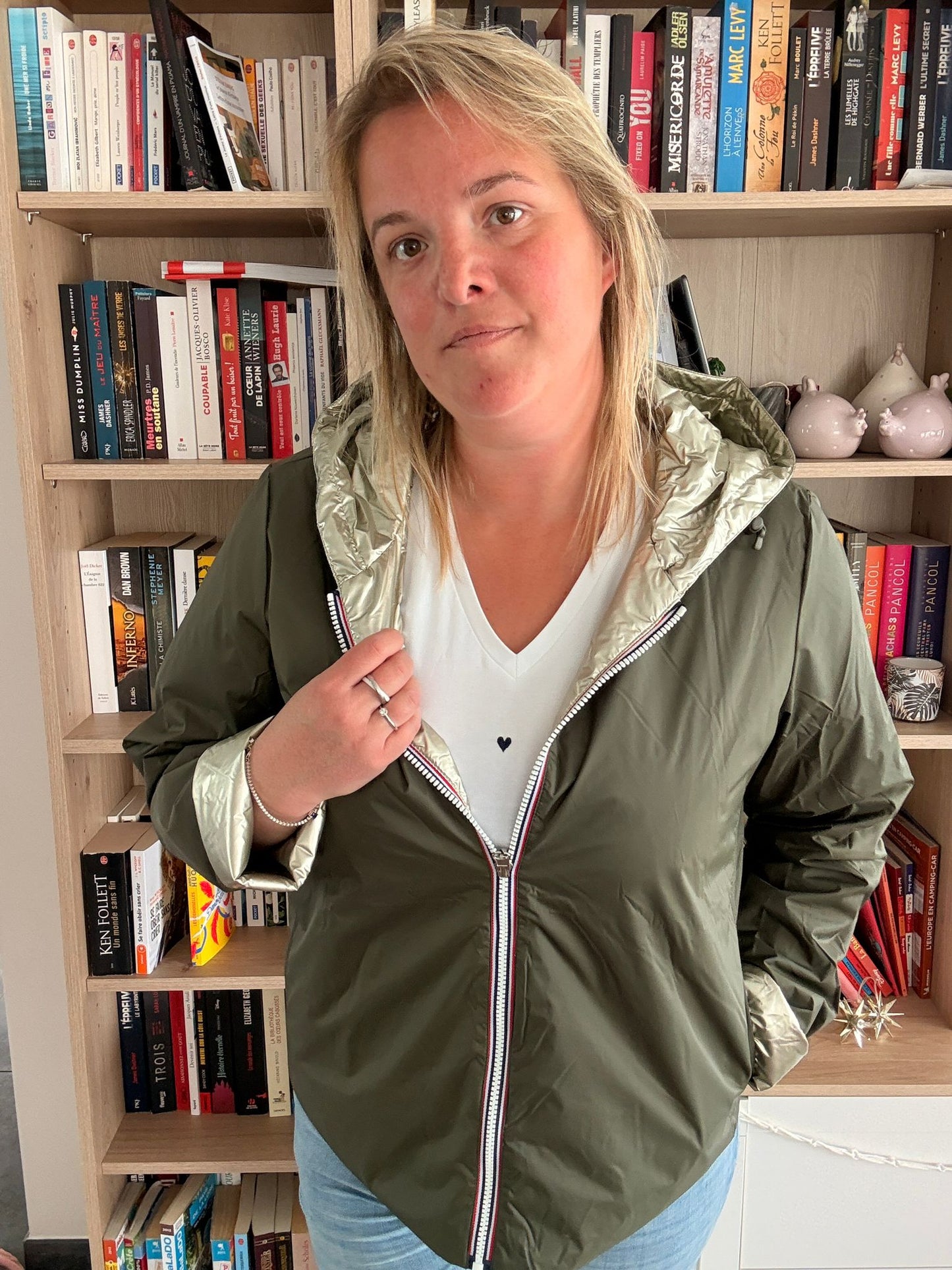 Imperméable réversible court kaki doré pour femme porté par une personne tenant la fermeture, en intérieur avec bibliothèque en arrière-plan.