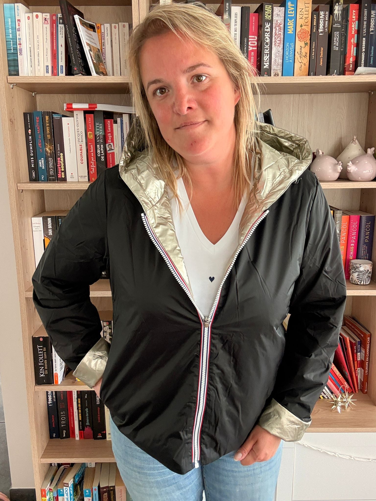 Imperméable réversible court noir doré femme Laura Jo Mi-Saison : femme posant devant bibliothèque, veste noire avec capuche dorée et ourlets dorés, zippé, jeans bleu.