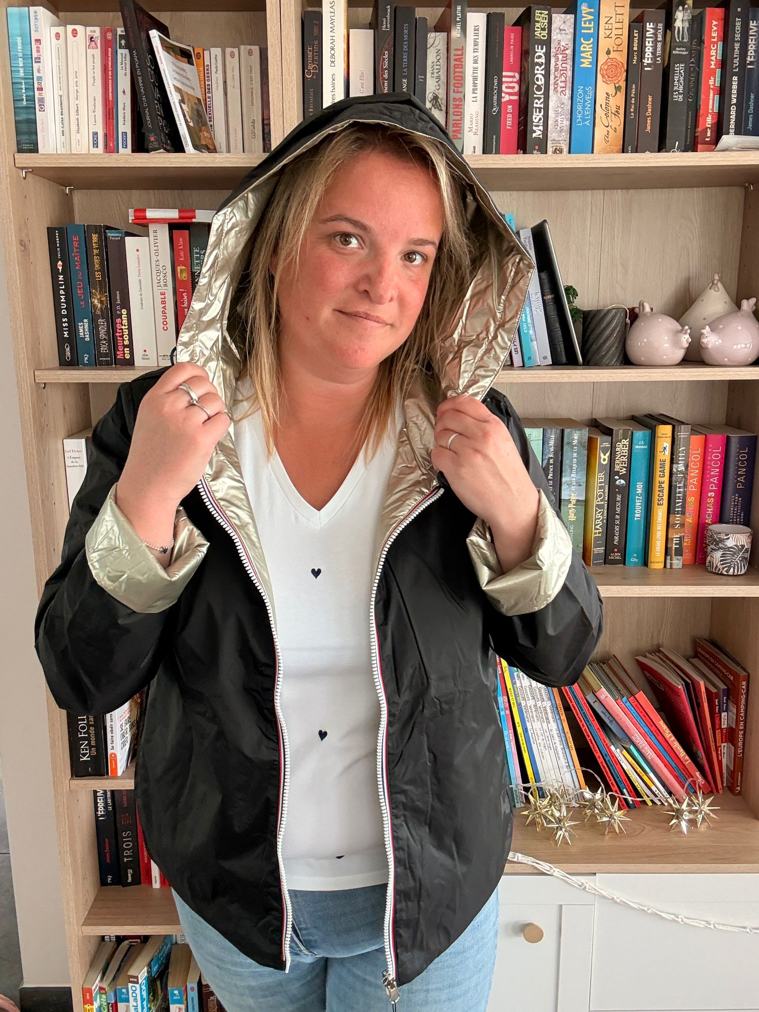 Imperméable réversible noir-or doré porté par une femme, capuche relevée, devant une bibliothèque remplie de livres.