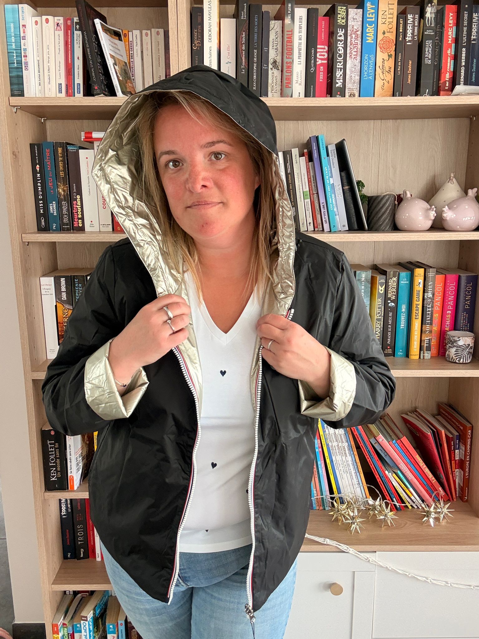 Imperméable réversible court noir doré femme, portée par une personne devant une étagère de livres, capuche relevée et doublure dorée visible.