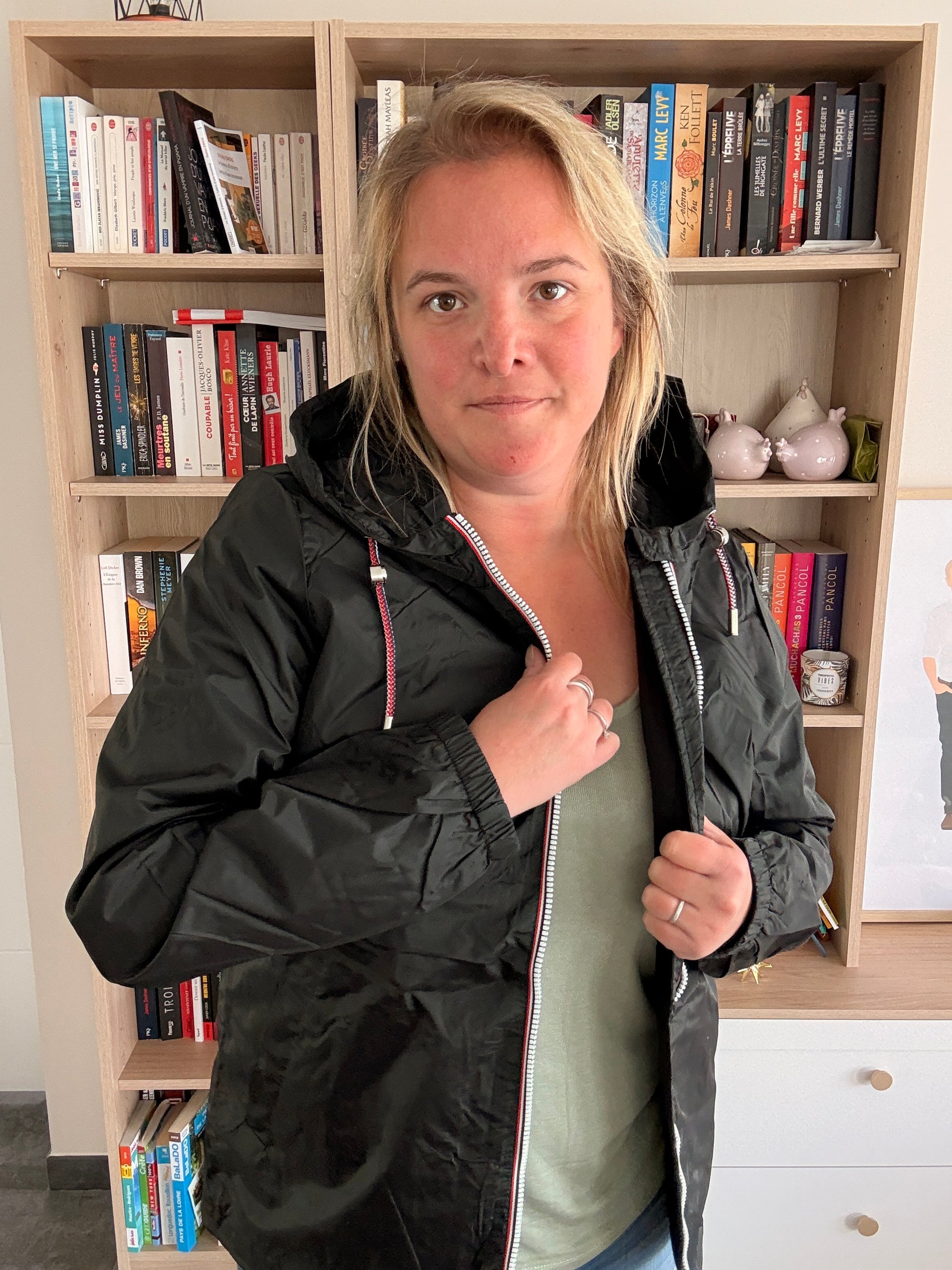 Femme de 3/4 portant l'imperméable court noir Laura Jo ouvert, main sur le zip bas, poches latérales visibles