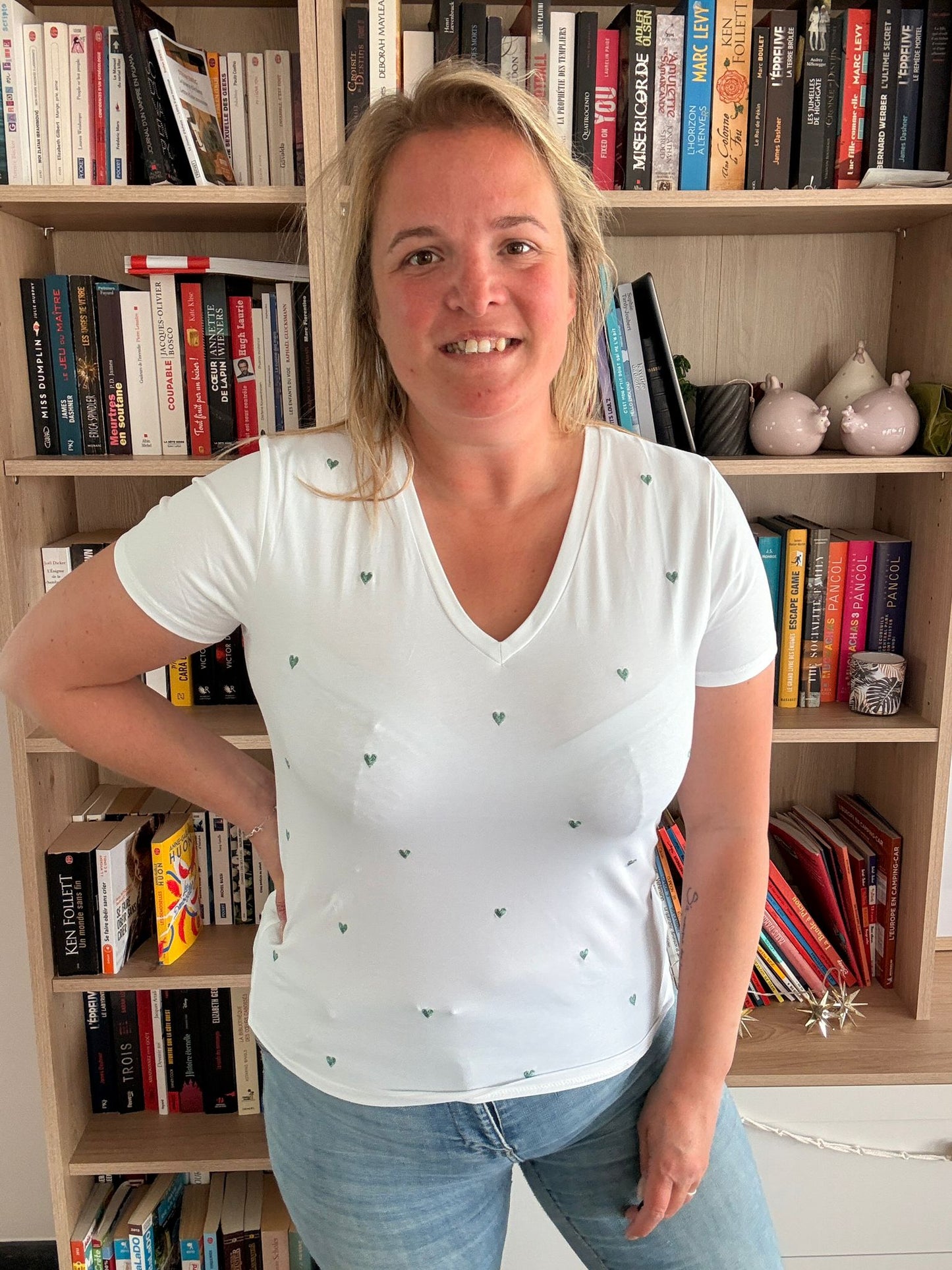 T-shirt blanc à col V orné de petits cœurs brodés verts porté par une femme devant une bibliothèque.
