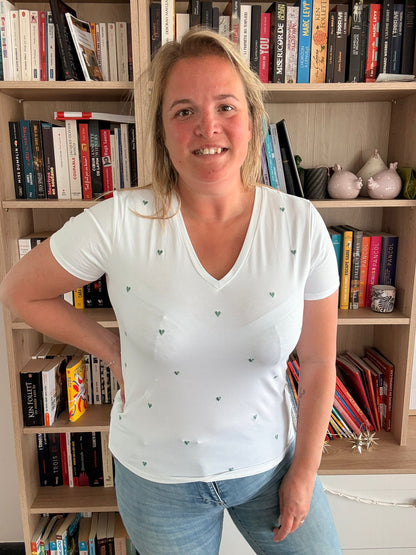 T-shirt blanc à col V orné de petits cœurs brodés verts porté par une femme devant une bibliothèque.