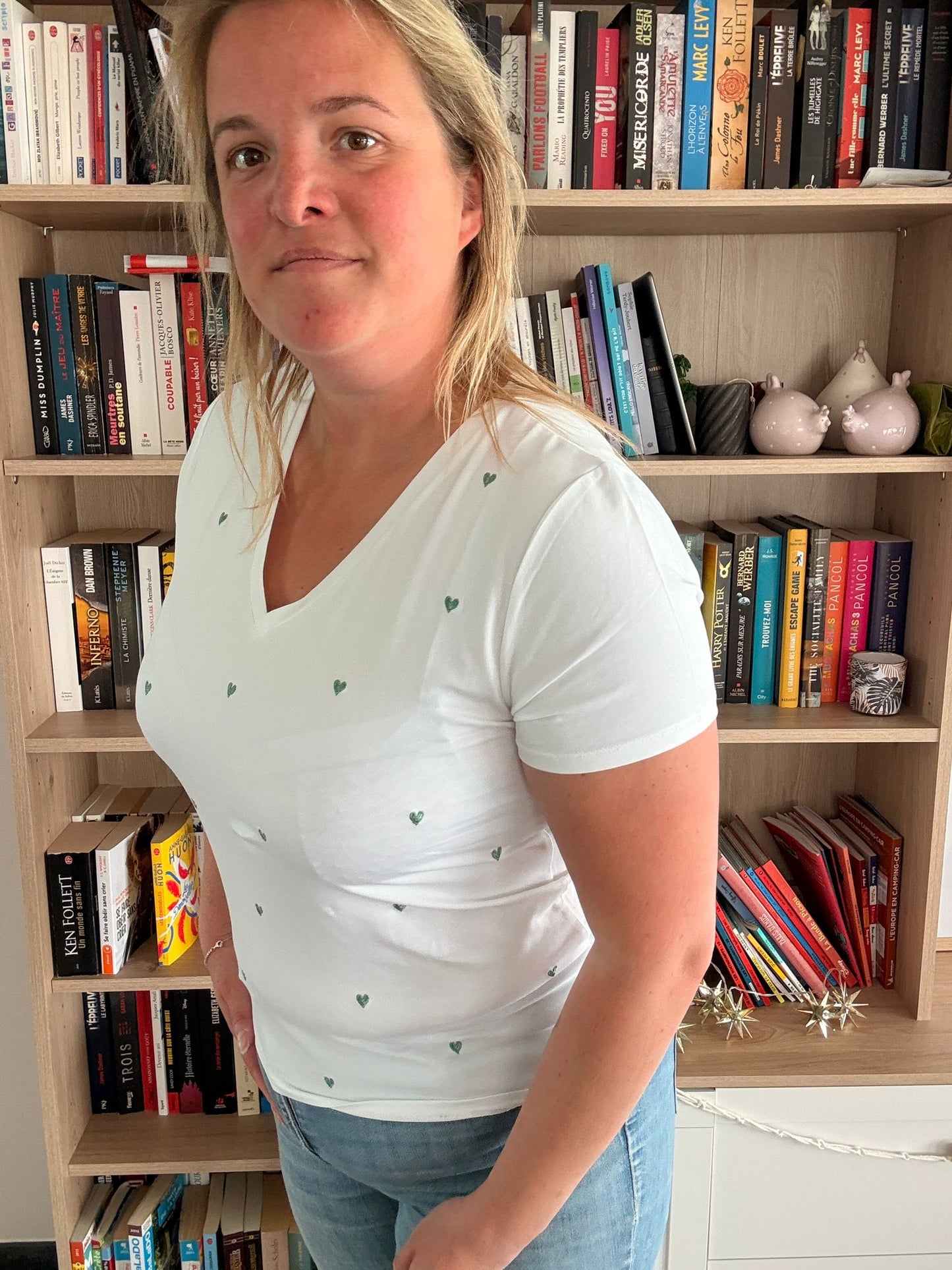 T-shirt blanc à petits cœurs verts brodés porté par une femme debout dans une bibliothèque.