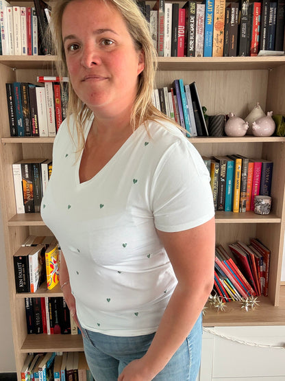 T-shirt blanc à petits cœurs verts brodés porté par une femme debout dans une bibliothèque.