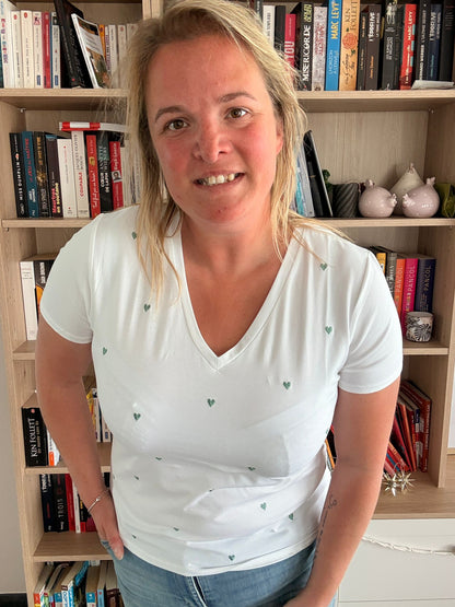 T-shirt blanc avec des cœurs verts brodés portée par une femme, en intérieur près d’une étagère de livres.