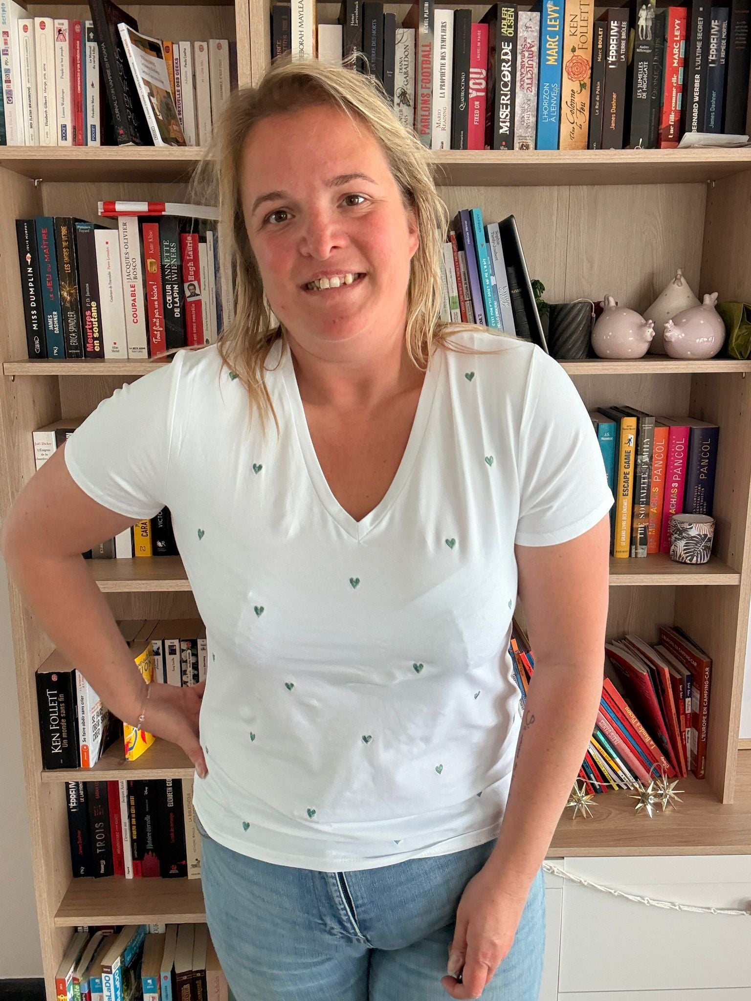 Homme ou femme portant un T-shirt blanc à petits cœurs verts, debout devant une étagère de livres, sourire détendu.