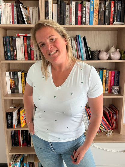 T-shirt blanc à cœurs brodés verts porté par une femme souriante devant une étagère de livres.