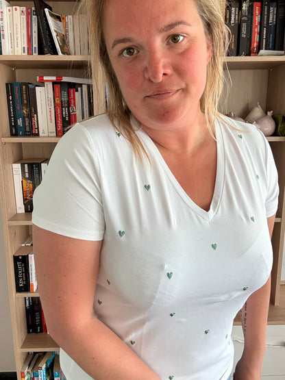 T-shirt blanc à col V, décoré de petits cœurs verts brodés, porté par une femme près d’une étagère de livres.