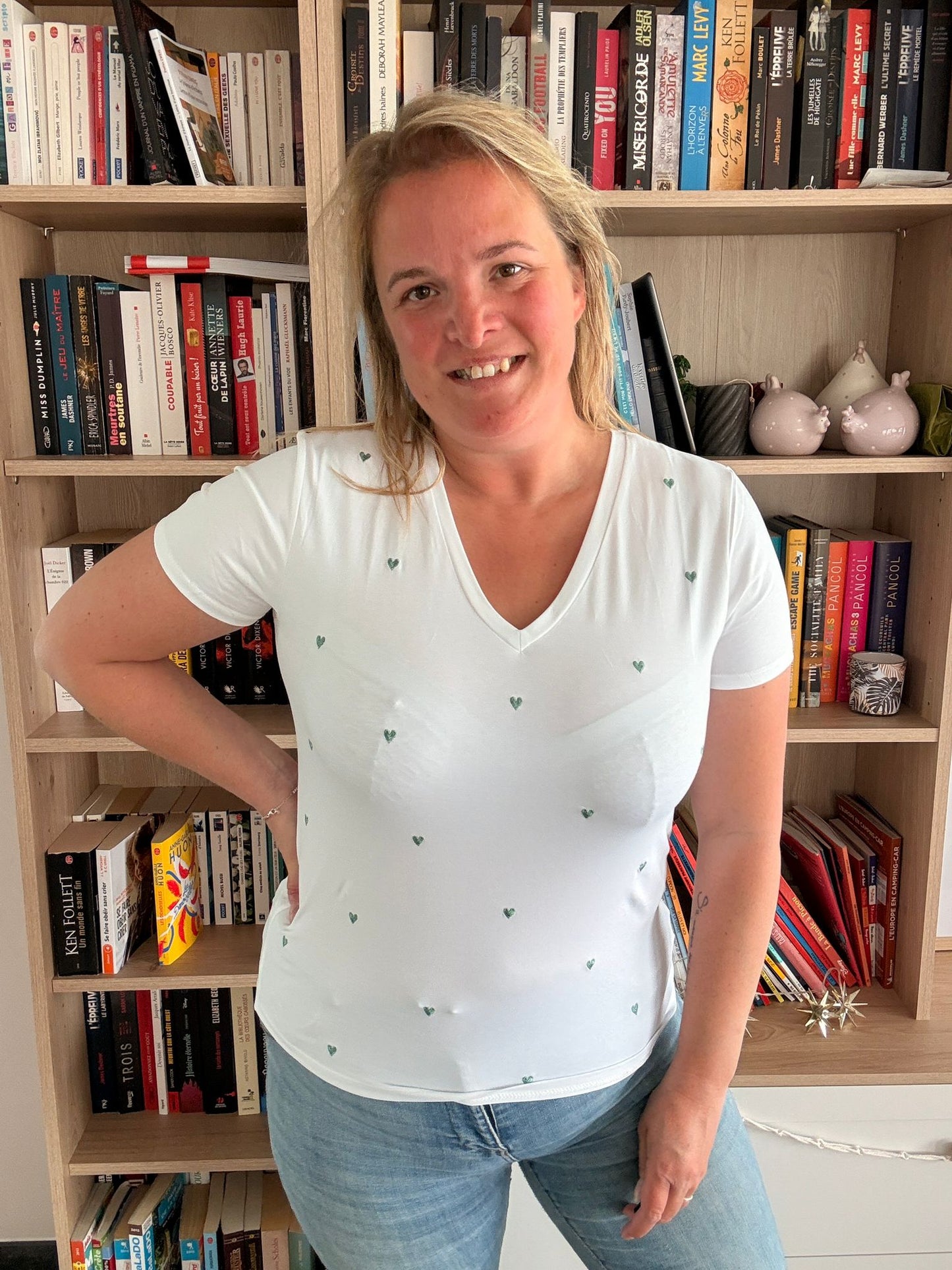 T-shirt blanc à cœur brodé vert porté par une femme devant une étagère de livres.