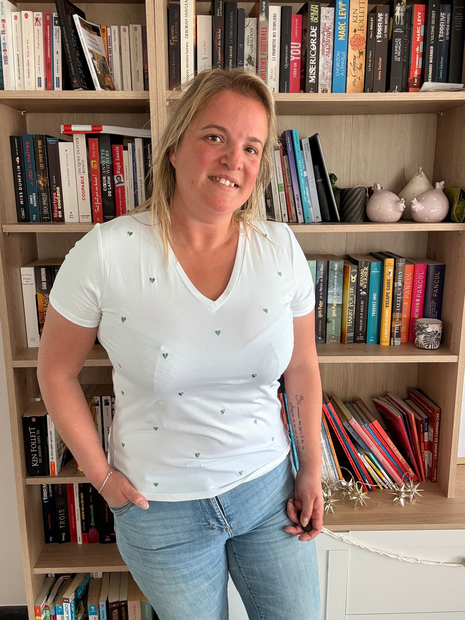 T-shirt blanc avec petits cœurs brodés verts porté par une femme souriante devant une bibliothèque.