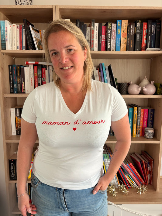 T-shirt blanc ajusté avec inscription « maman d'amour » et petit cœur rouge, porté par une femme près d'une étagère de livres.