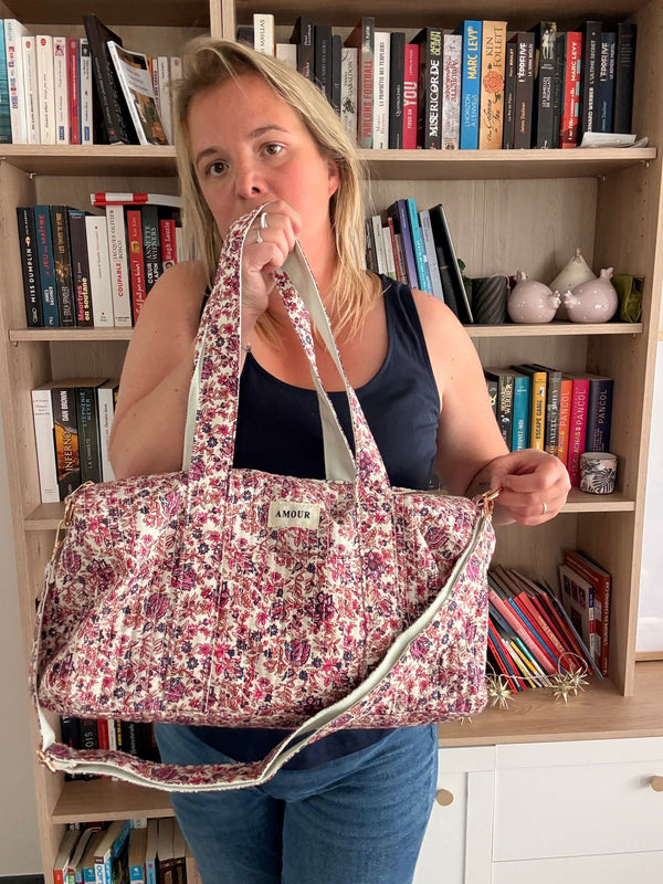 Sac polochon “Amour” en coton matelassé fleuri rose et bordeaux, porté par une femme en intérieur, bibliothèque en arrière-plan.