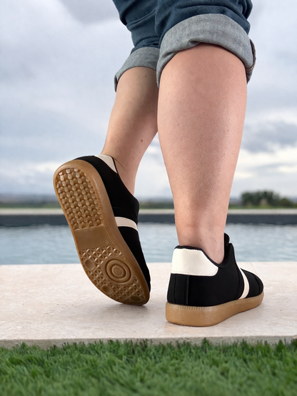 Vue arrière de baskets basses noires bande blanche femme style sneakers campus, semelle gomme caramel, portées près d'une piscine