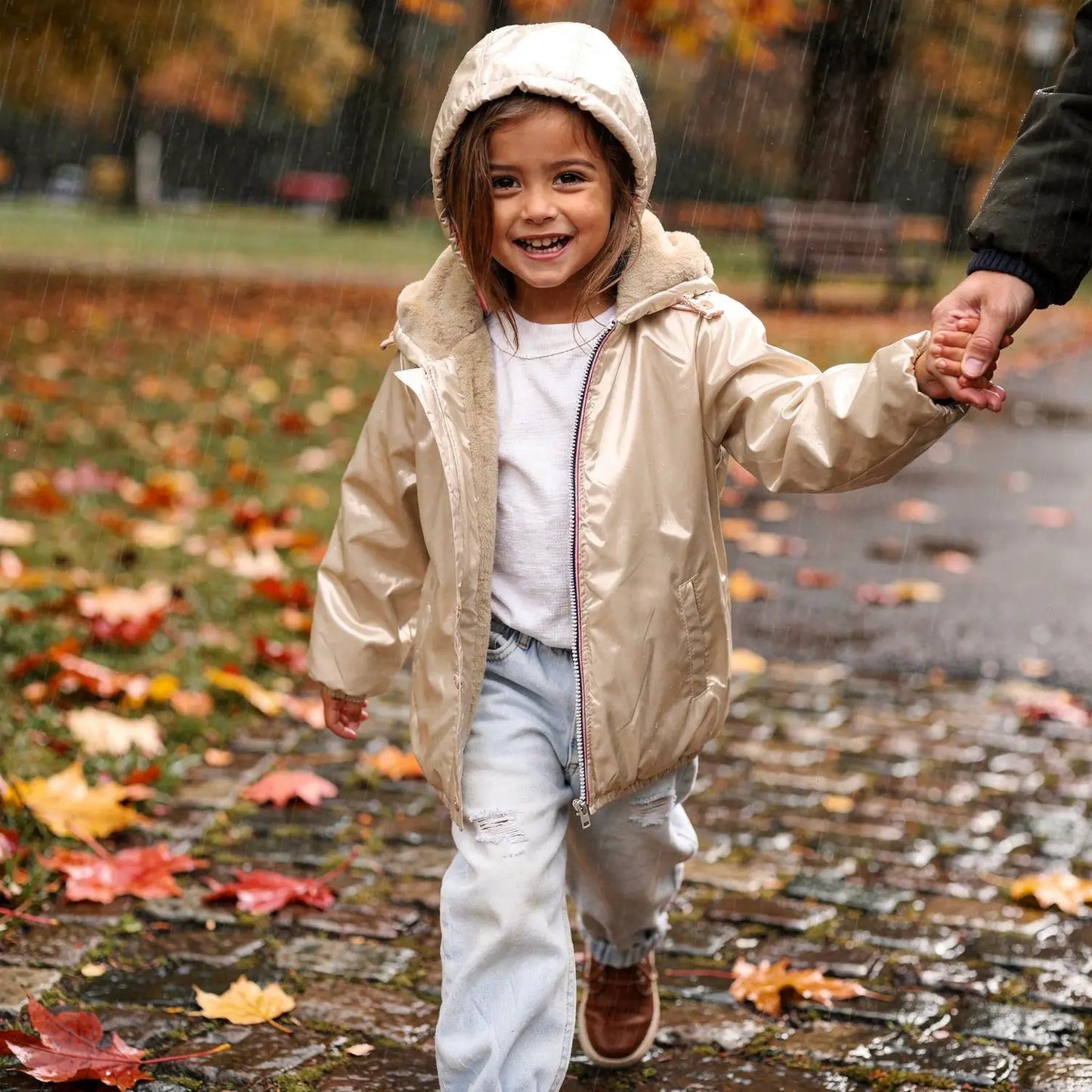 Enfant fille portant un imperméable doré doublé avec capuche, veste courte, kaki clair, marchant sous la pluie avec une main tenue par un adulte.