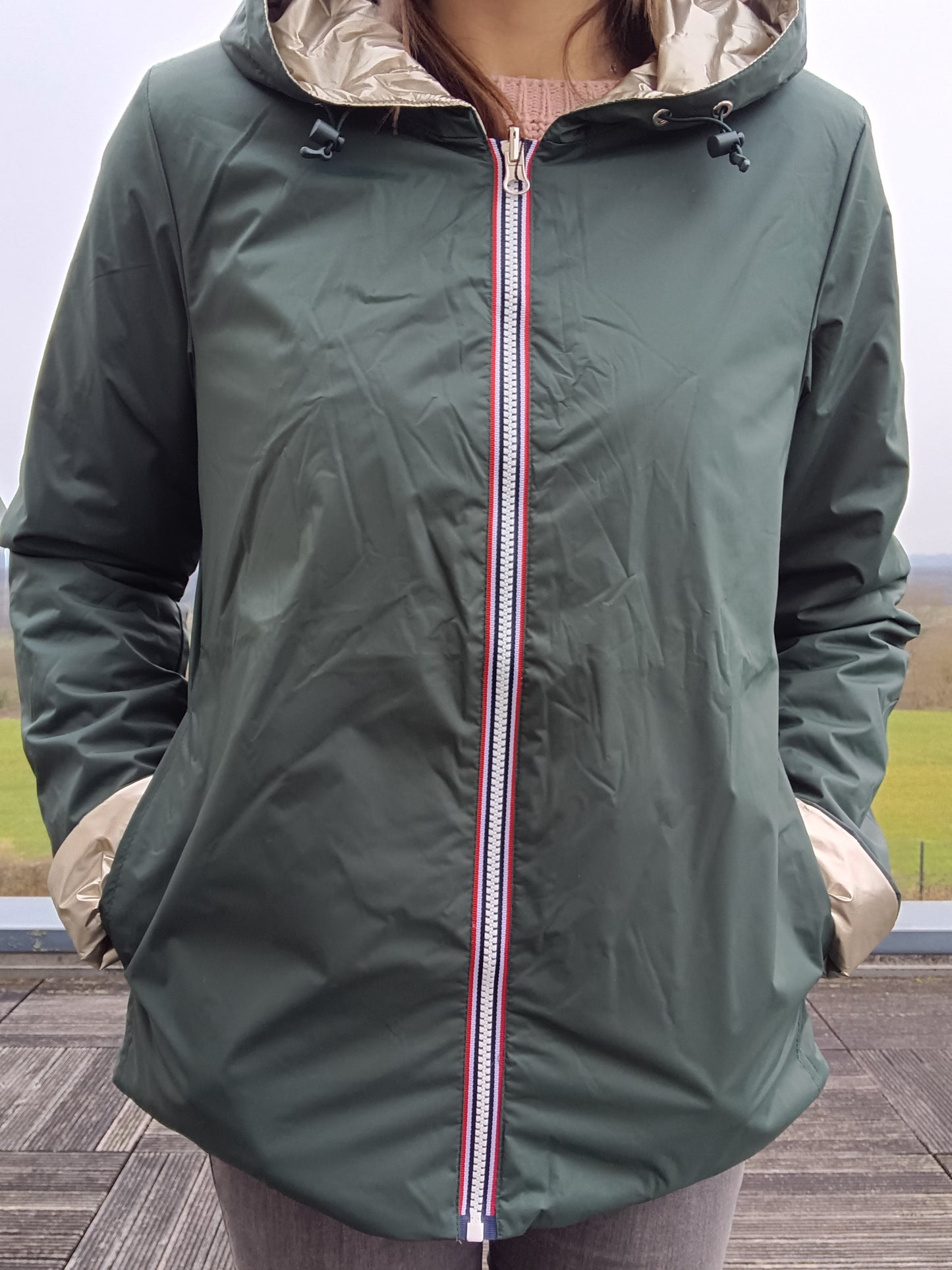 Veste imperméable réversible verte, fermeture éclair centrale avec bordure tricolore, capuche beige intérieur et manches retroussées.
