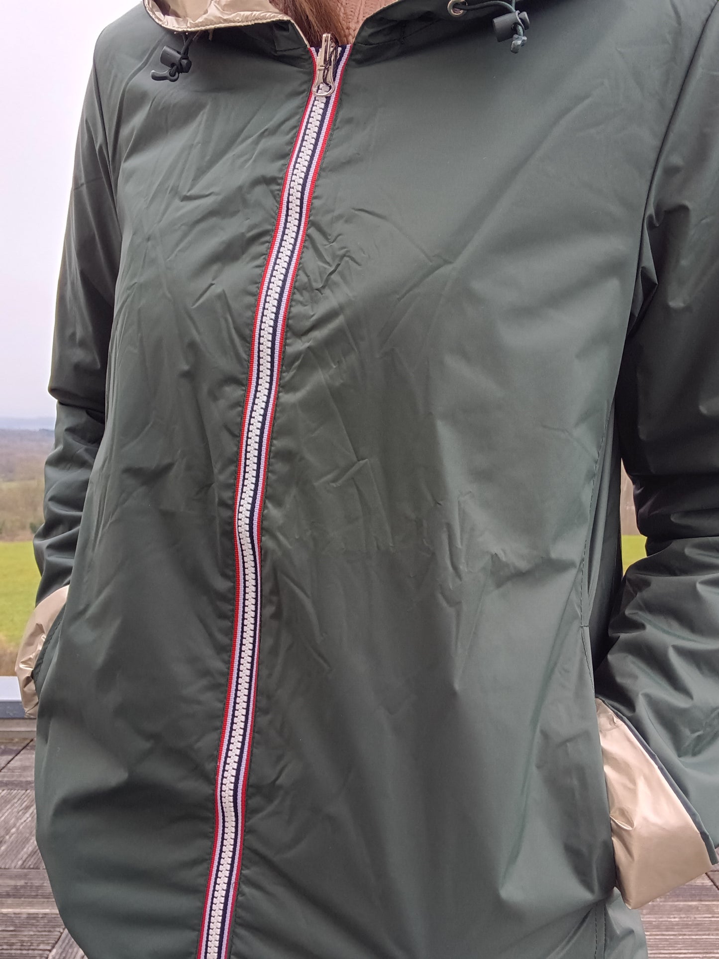 Veste imperméable réversible vert forêt / doré, coupe courte avec capuche, fermeture éclair centrale à rubans tricolores, doublure sombre, manches pliées, portée en plein air. Laura Jo