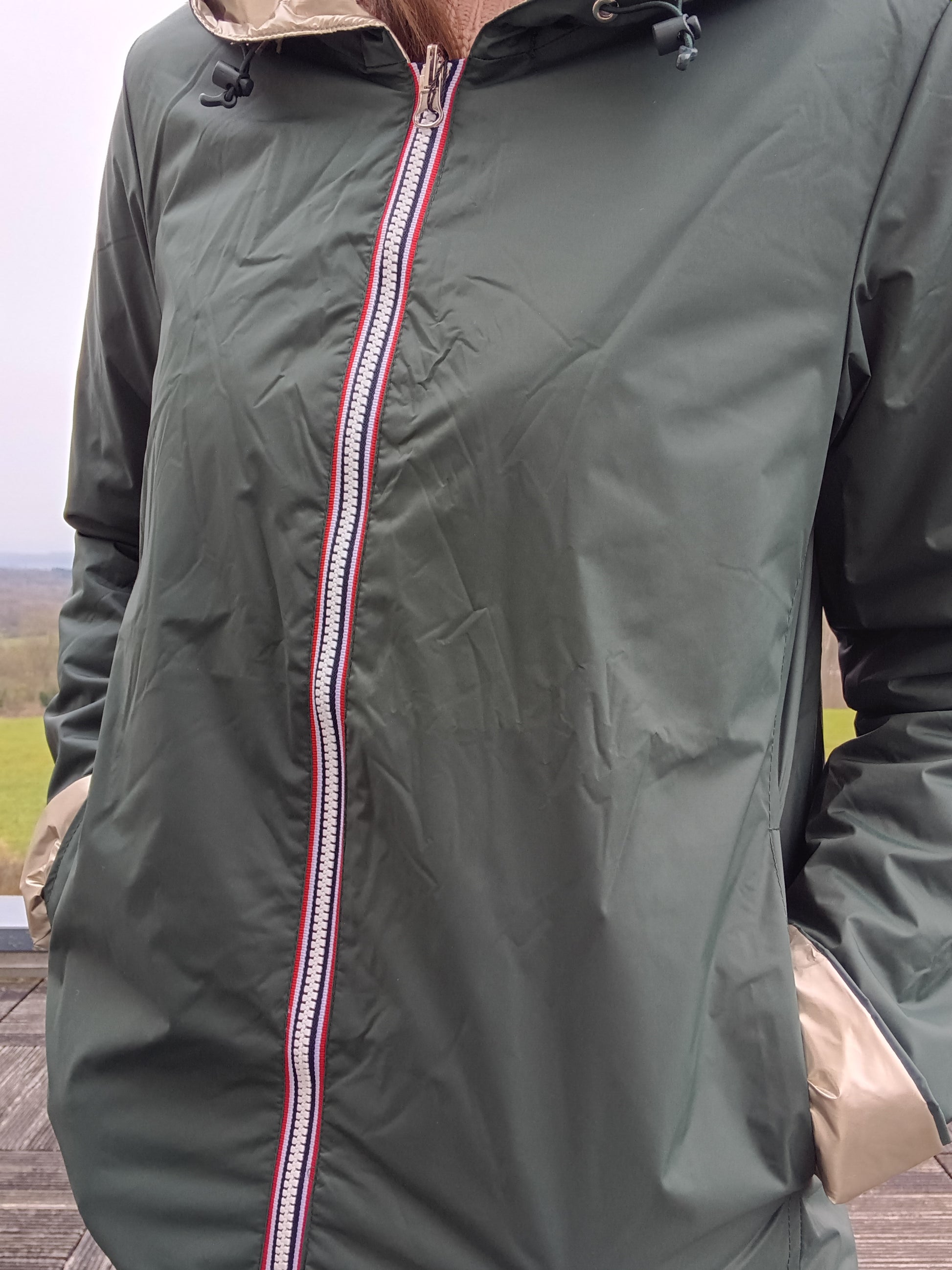 Veste imperméable réversible vert forêt / doré, coupe courte avec capuche, fermeture éclair centrale à rubans tricolores, doublure sombre, manches pliées, portée en plein air. Laura Jo