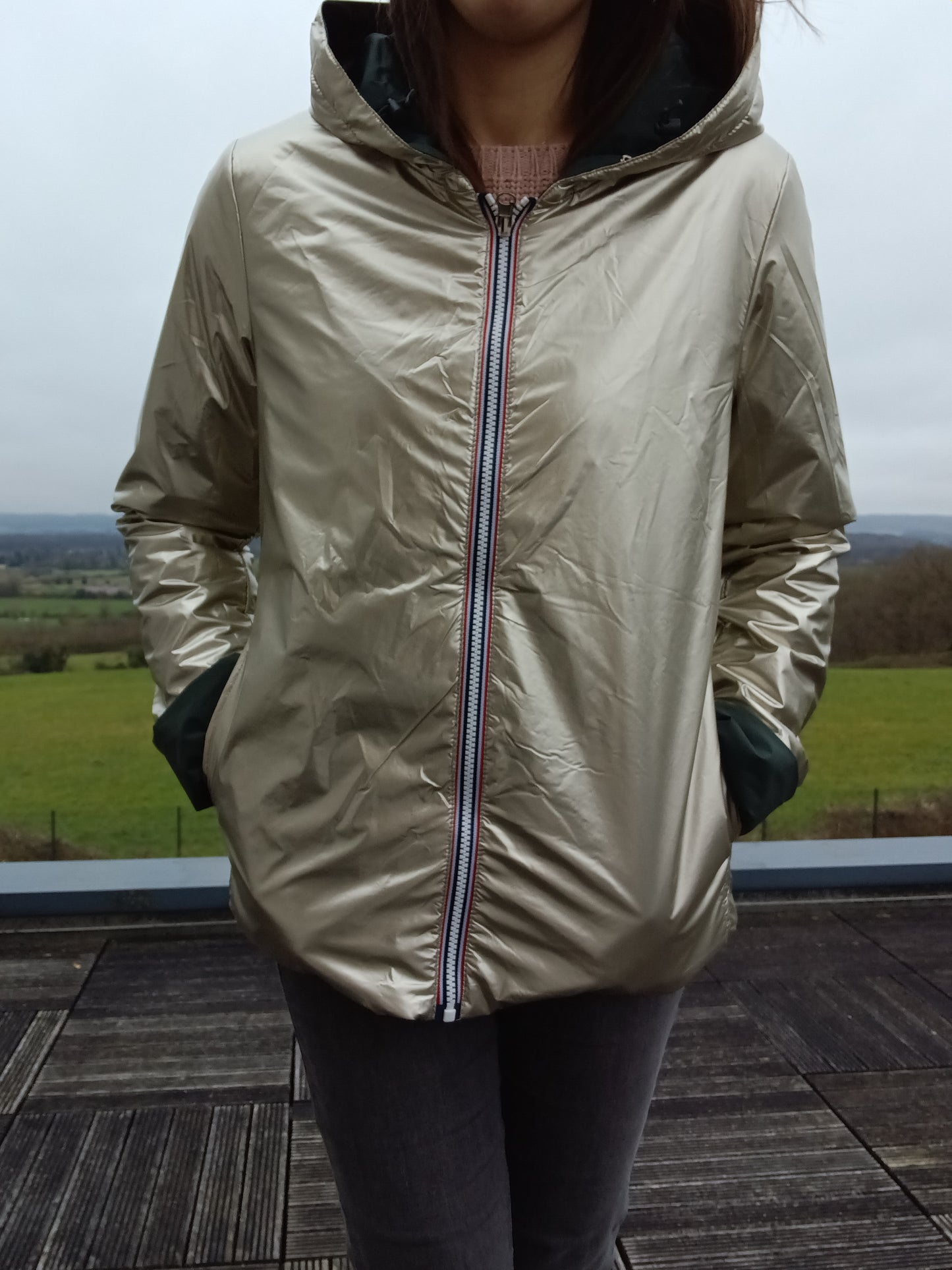 Veste imperméable réversible beige clair, coupe courte avec capuche, fermeture éclair centrale à rubans tricolores, doublure sombre, manches pliées, portée en plein air. Product: Laura Jo - Imperméable réversible court garnissage ouate mi-saison vert forêt / doré