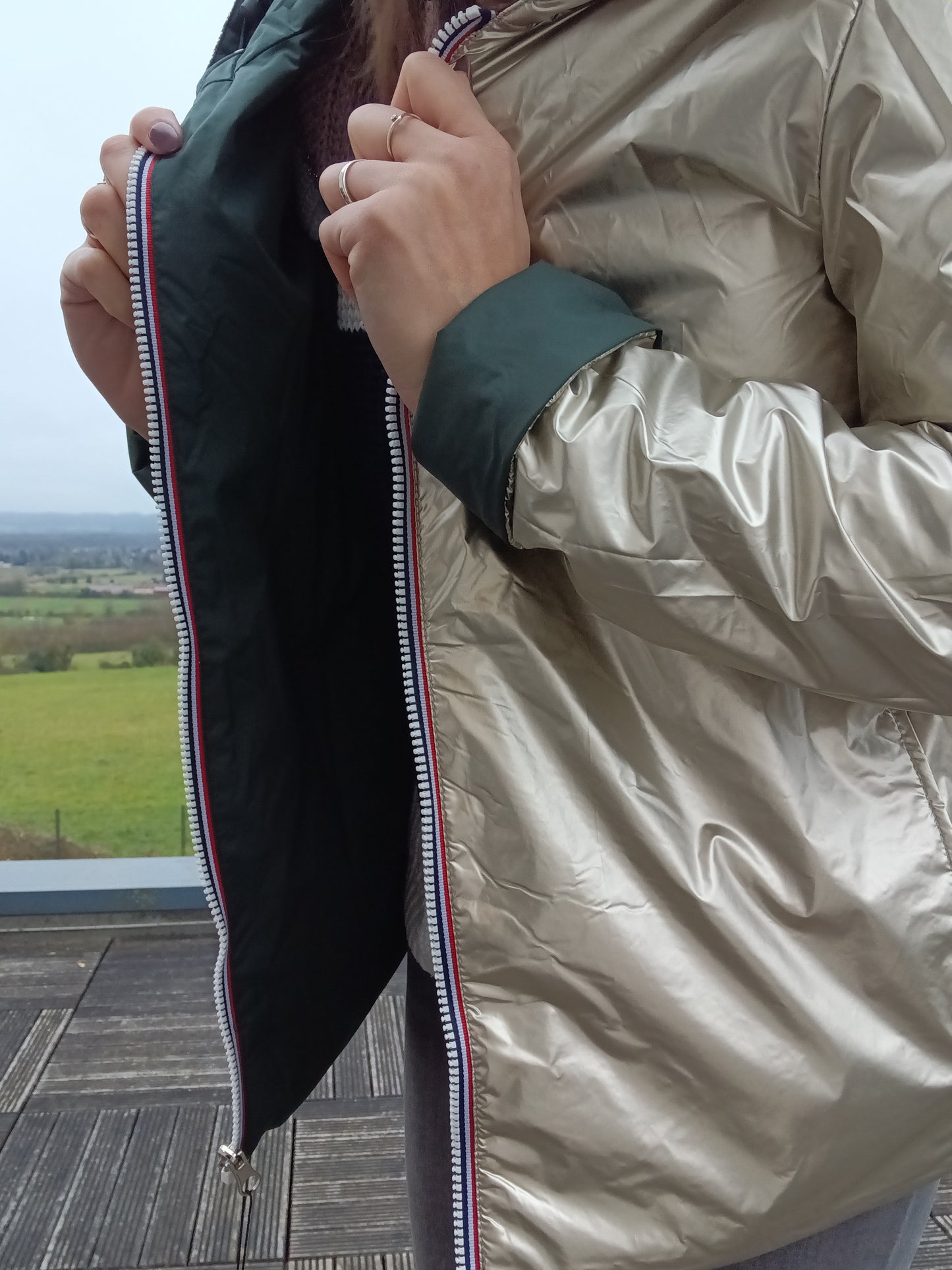 Veste imperméable réversible doré / vert forêt, coupe courte avec capuche, fermeture éclair centrale à rubans tricolores, manches pliées, portée en plein air. Laura Jo