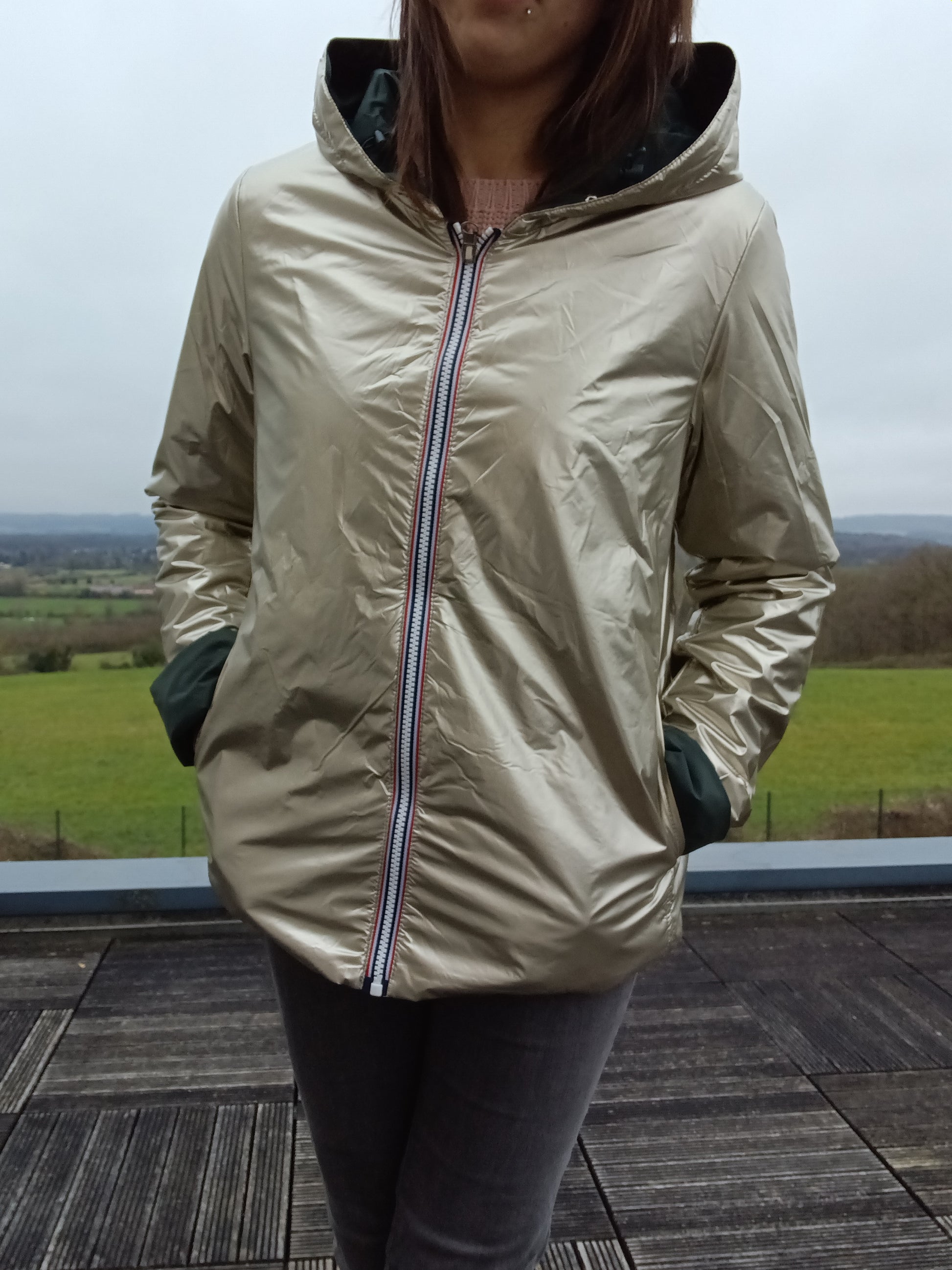 Veste imperméable réversible doré, coupe courte avec capuche, fermeture éclair centrale à rubans tricolores, doublure sombre, manches pliées, portée en plein air. Laura Jo