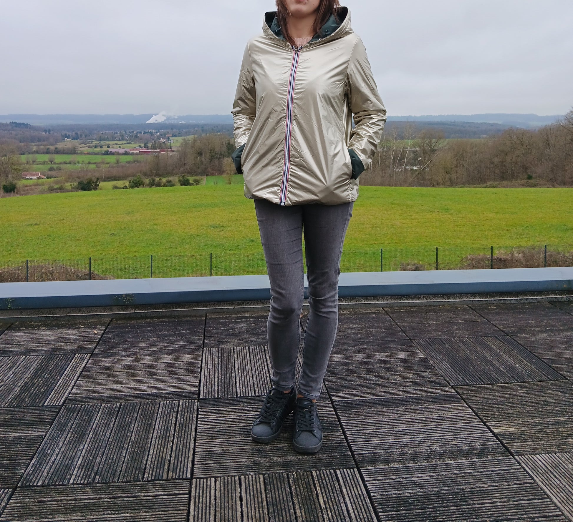 Veste imperméable réversible doré, coupe courte avec capuche, fermeture éclair centrale à rubans tricolores, doublure sombre, manches pliées, portée en plein air. Laura Jo