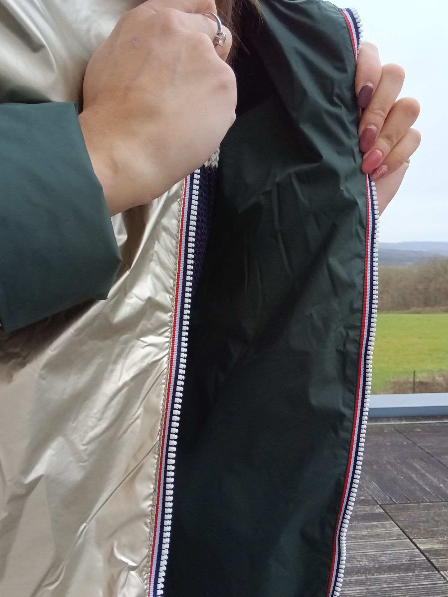 Veste imperméable réversible beige clair, coupe courte avec capuche, fermeture éclair centrale à rubans tricolores, doublure sombre, manches pliées, portée en plein air. Laura Jo - Imperméable réversible court mi-saison vert forêt / doré